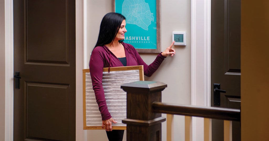 woman programming a new smart thermostat while holding a new furnace filter replacement